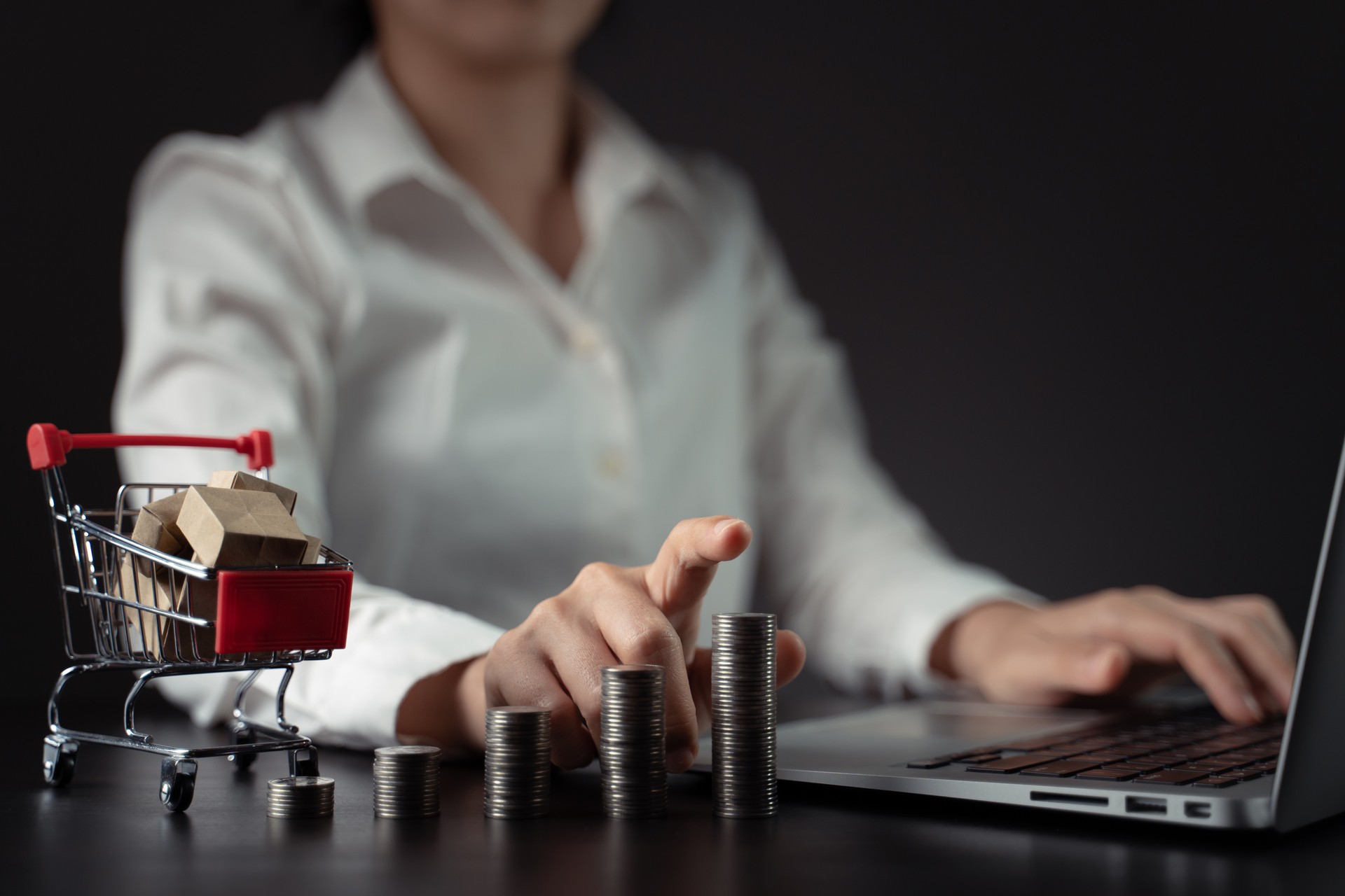 Close up shot of woman typing on laptop with mini shopping cart and coins stack.